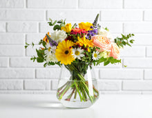 A bright and elegant garden-style bouquet in a glass vase, featuring roses, hydrangeas, seasonal blooms, and lush greenery in a cheerful, mixed-color palette.