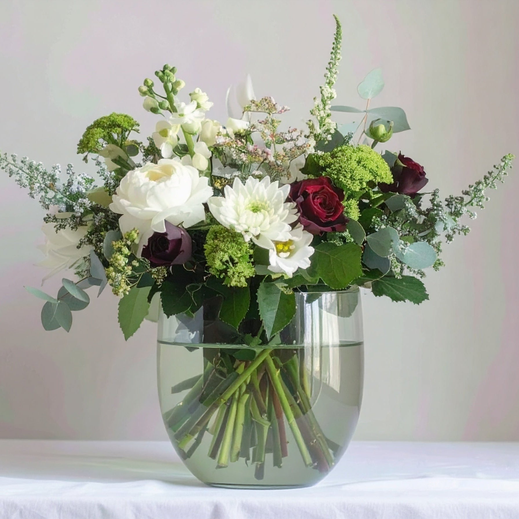 A sophisticated floral arrangement featuring white blooms, vibrant roses, and lush greenery, elegantly designed in a natural, garden-inspired style.