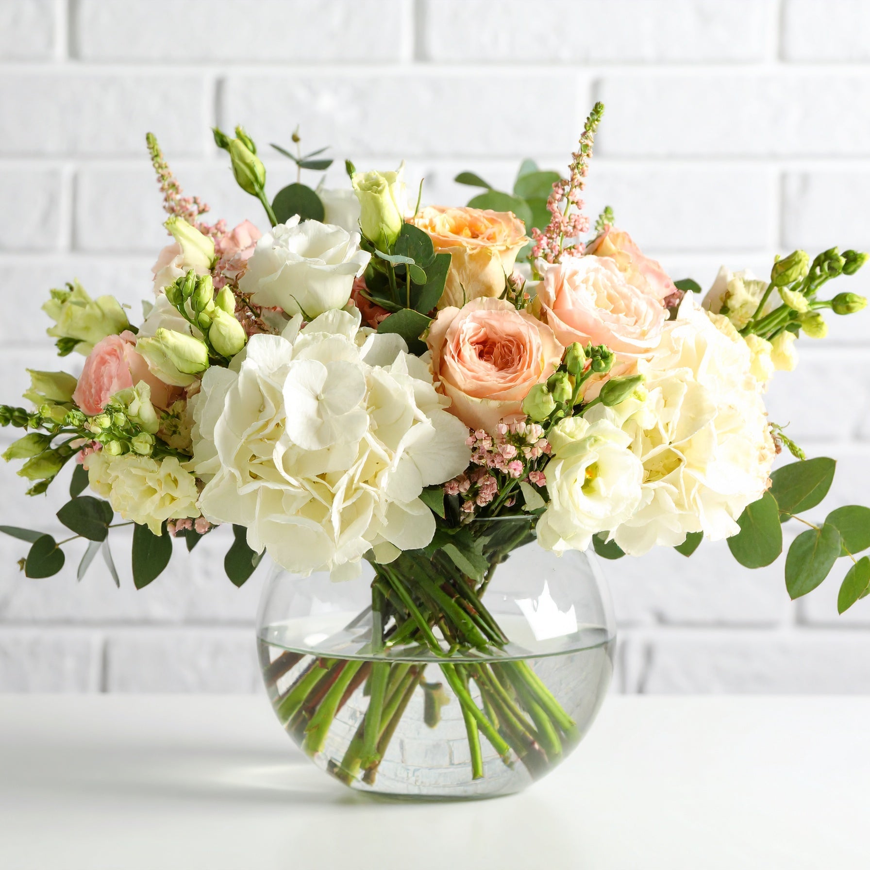 A pastel floral arrangement in a clear glass vase, featuring soft-hued blooms like blush, lavender, peach, and cream flowers, accented with fresh greenery. 
